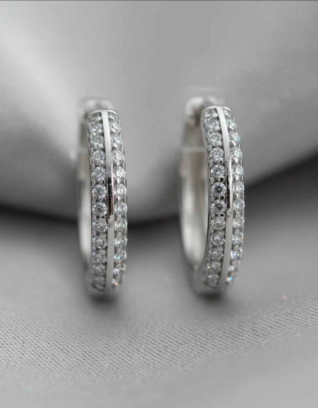 Par de pendientes de aro de plata con pequeñas piedras preciosas transparentes sobre fondo gris.