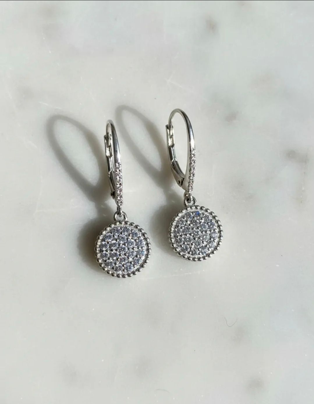 Par de pendientes colgantes de plata con colgantes redondos pavé sobre una superficie de mármol claro.