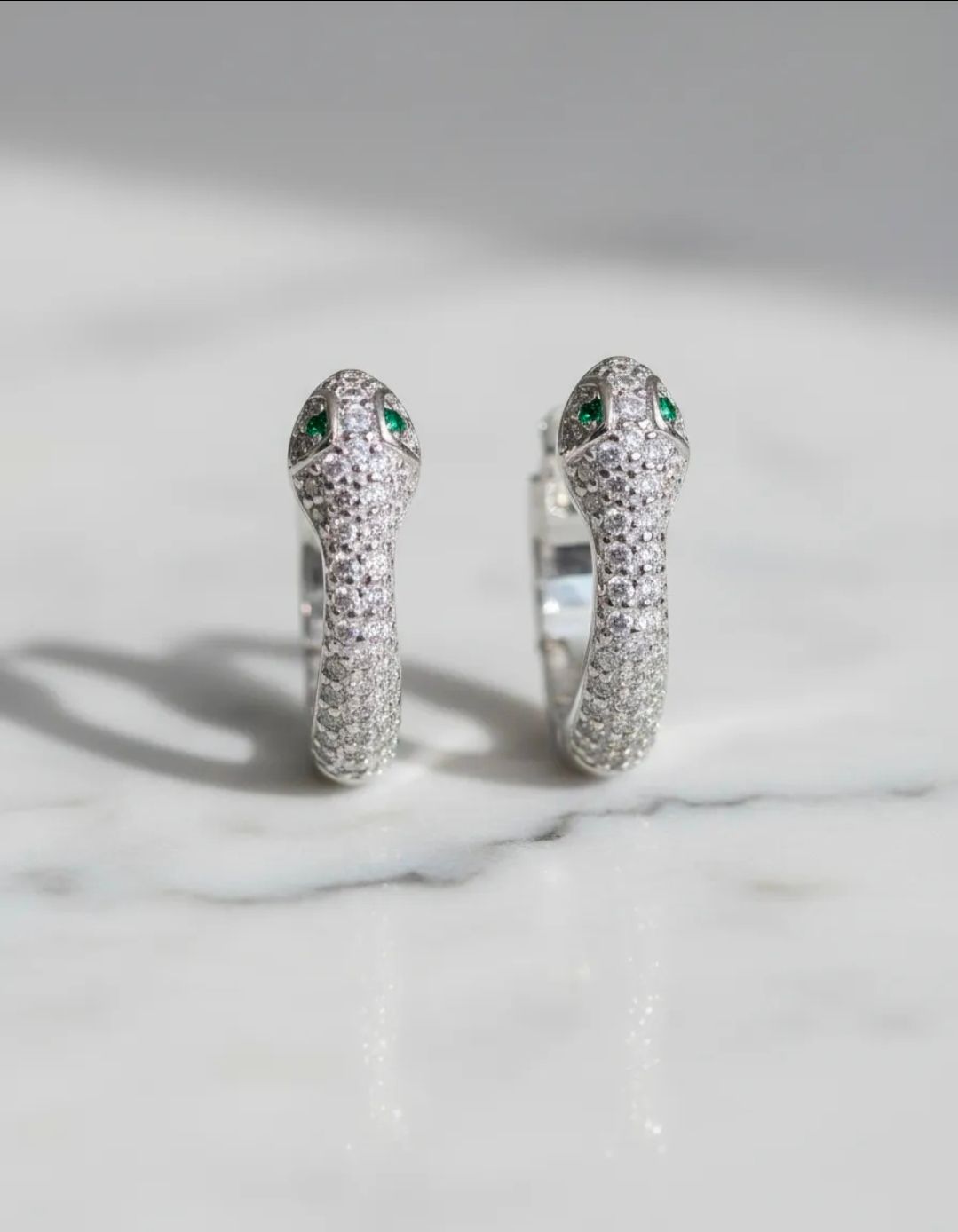 Par de pendientes de aro plateados con forma de serpiente y ojos de piedras preciosas verdes sobre una superficie de mármol.