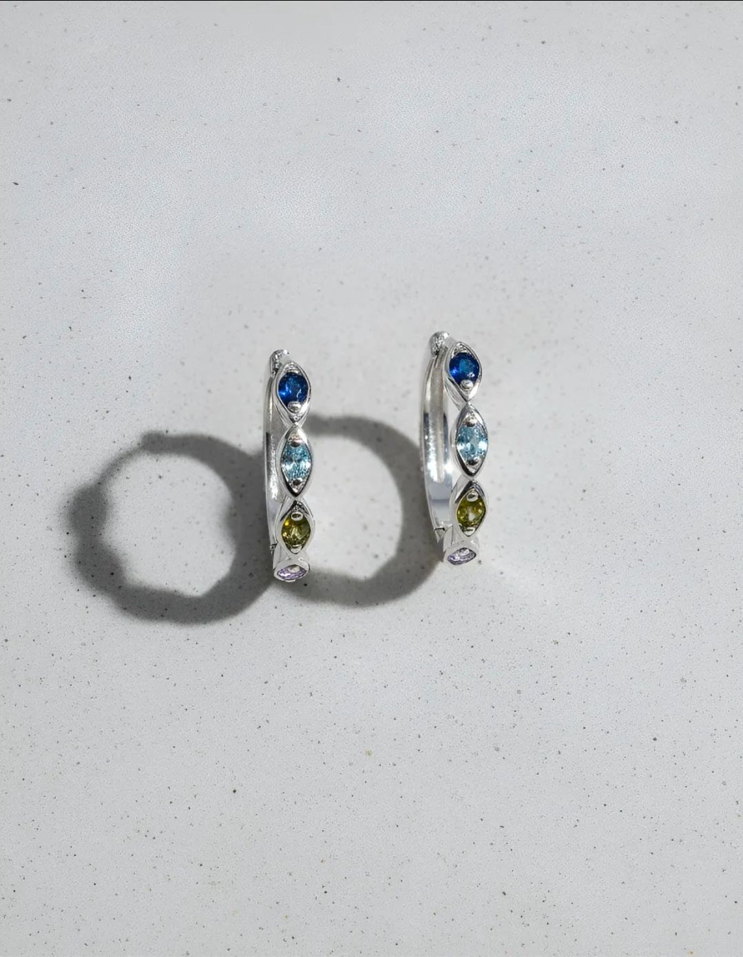 Dos pendientes de aro de plata con piedras preciosas azules, verdes y moradas sobre un fondo claro.