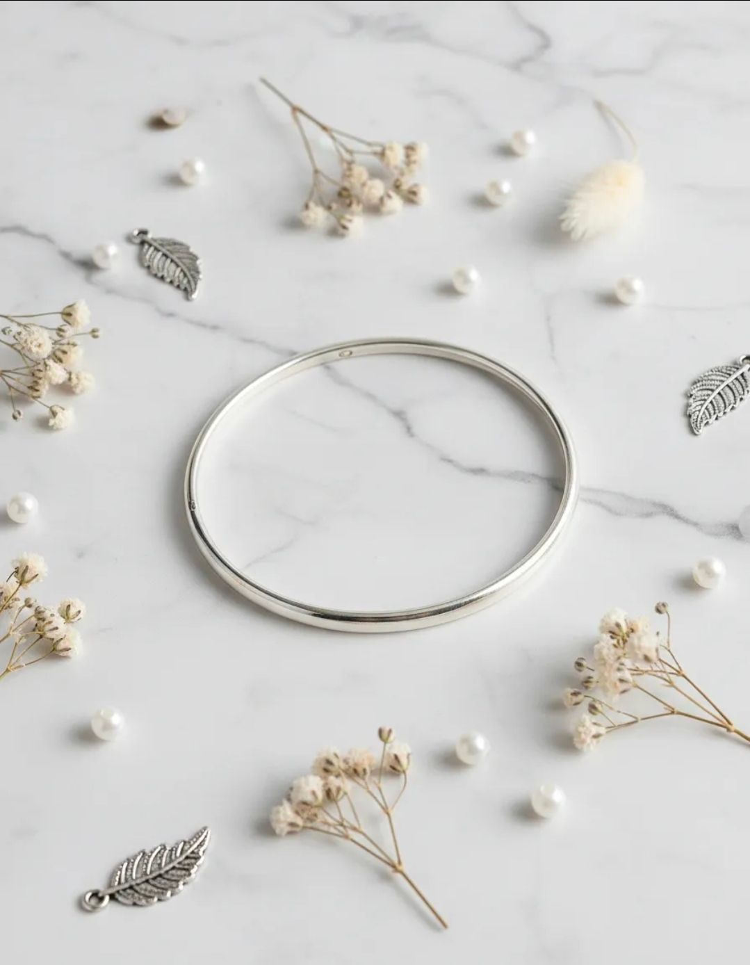 Brazalete de plata sobre mármol blanco con flores secas y cuentas que imitan perlas.