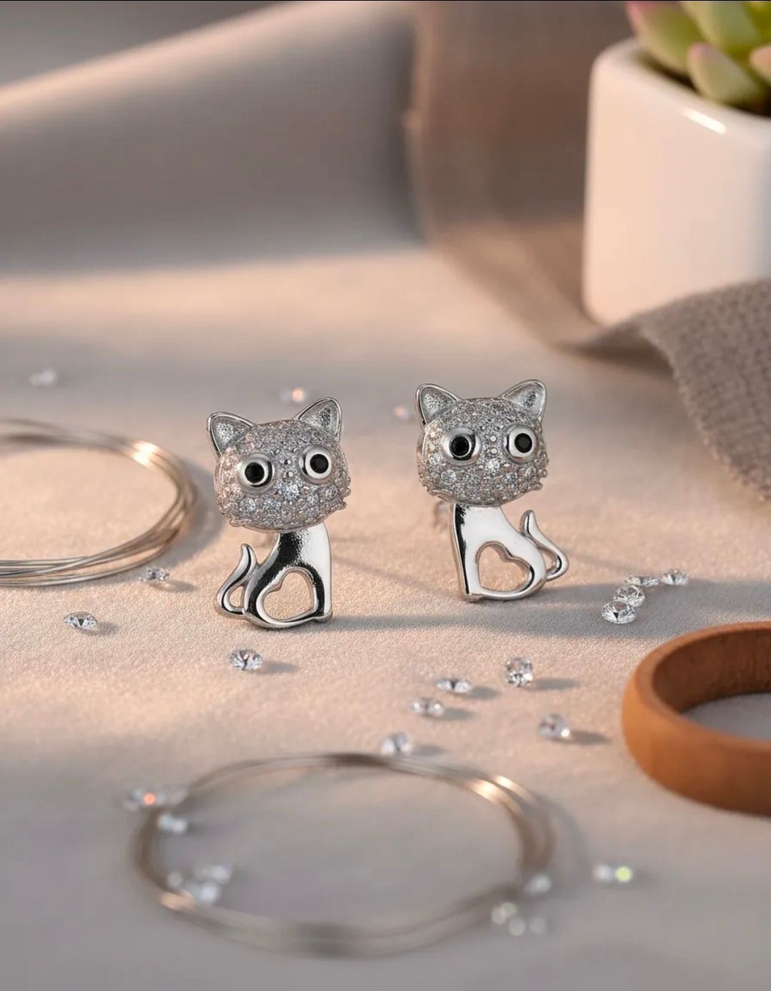 Un par de pendientes plateados con forma de gato sobre una mesa suave con anillos y una planta en maceta al fondo.