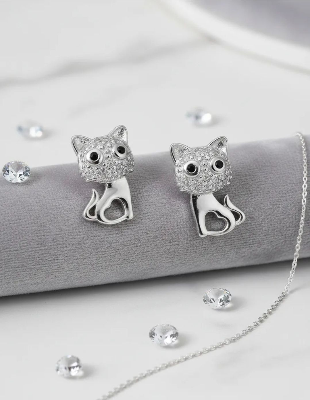 Un par de pendientes plateados con forma de gato sobre una mesa suave con anillos y una planta en maceta al fondo.