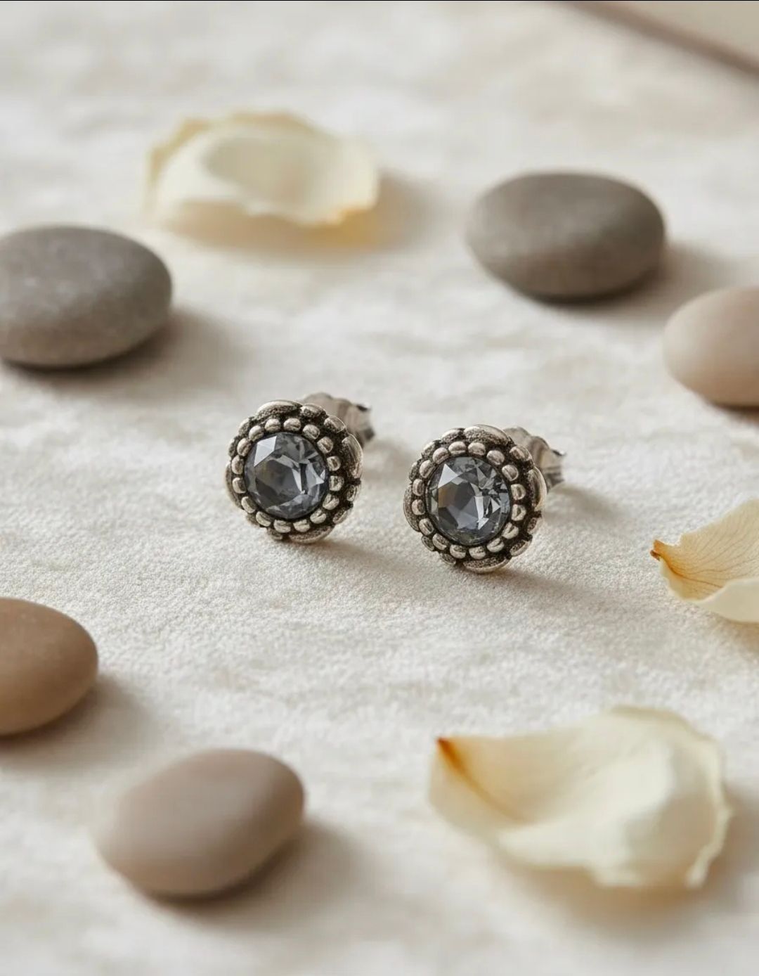 Par de pendientes de plata con forma de flor sobre una superficie beige claro con guijarros y pétalos blancos.