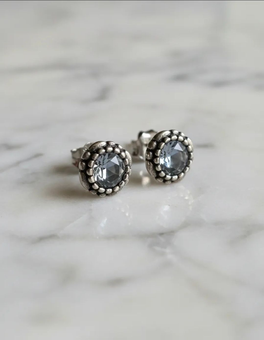 Par de pendientes de plata con forma de flor sobre una superficie beige claro con guijarros y pétalos blancos.