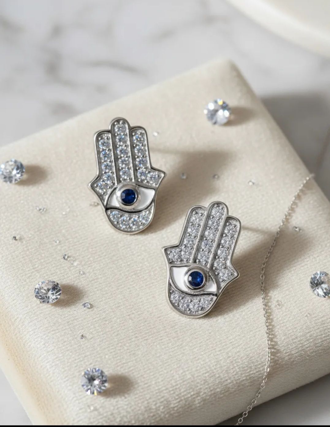 Pendientes de plata con forma de mano de Fátima y centros azules sobre una base color crema, rodeados de pequeñas gemas.