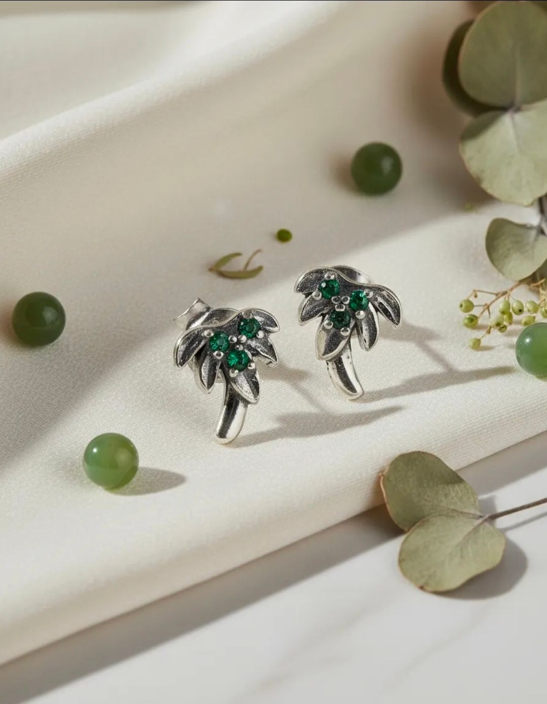 Pendientes de plata con forma de palmera y piedras verdes sobre una superficie de mármol blanco.