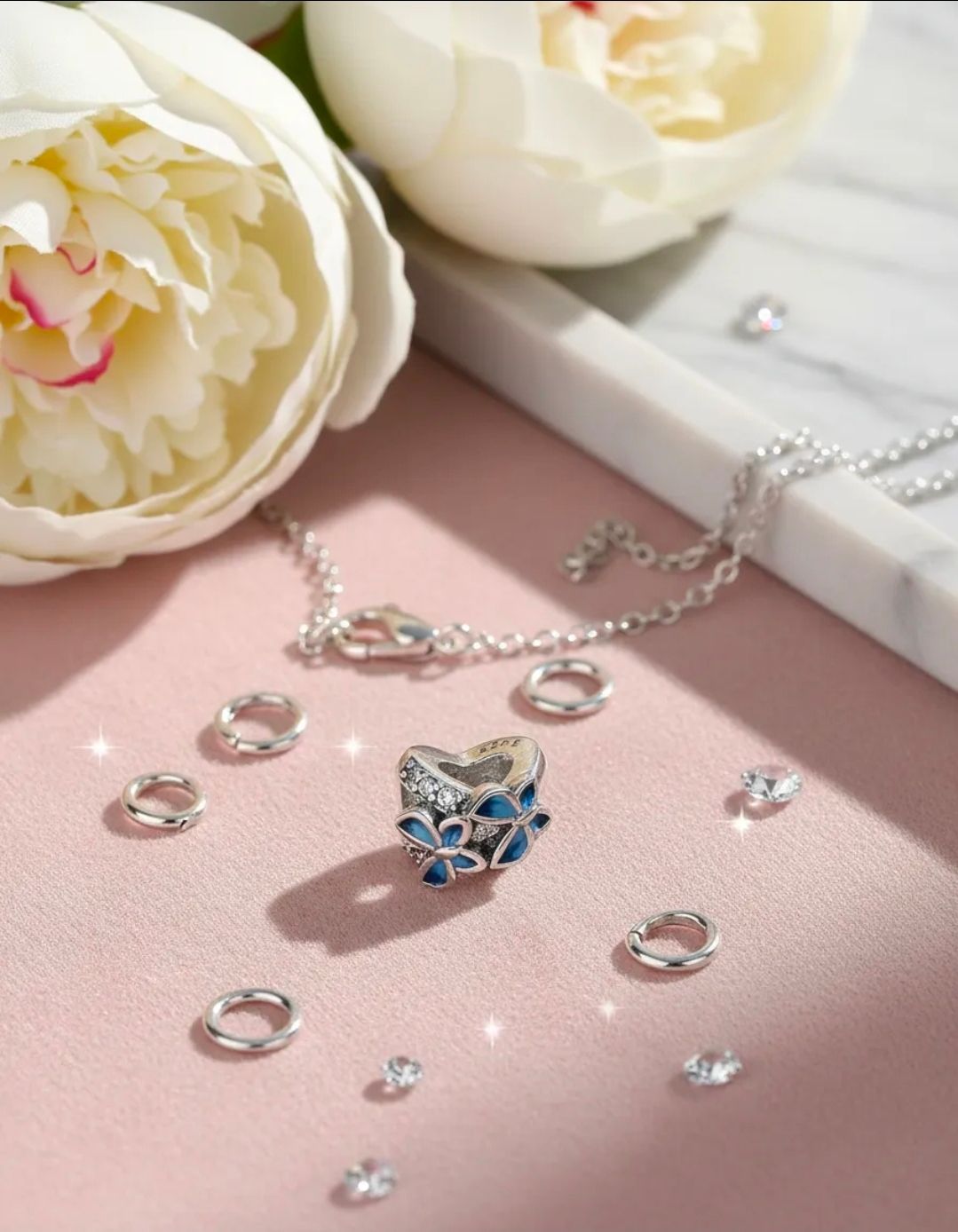 Joya de plata en forma de corazón con piedras azules sobre una superficie rosa, rodeada de perlas y flores blancas.