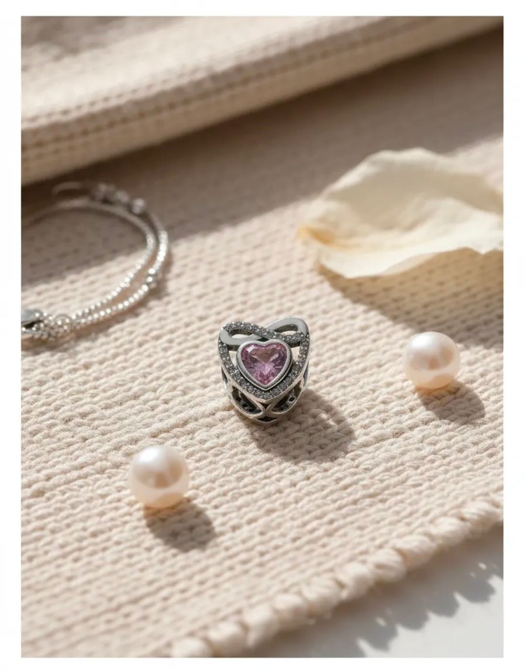 Anillo de plata en forma de corazón con piedra rosa, pendientes de perlas y una delicada cadena sobre tela beige.