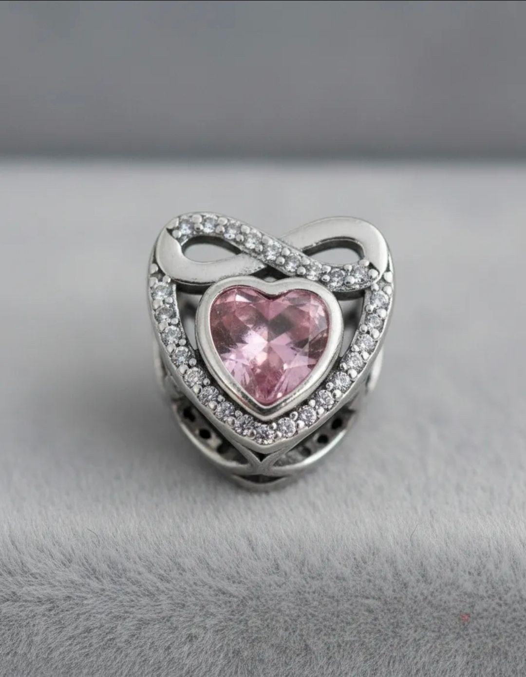 Anillo de plata en forma de corazón con piedra rosa, pendientes de perlas y una delicada cadena sobre tela beige.