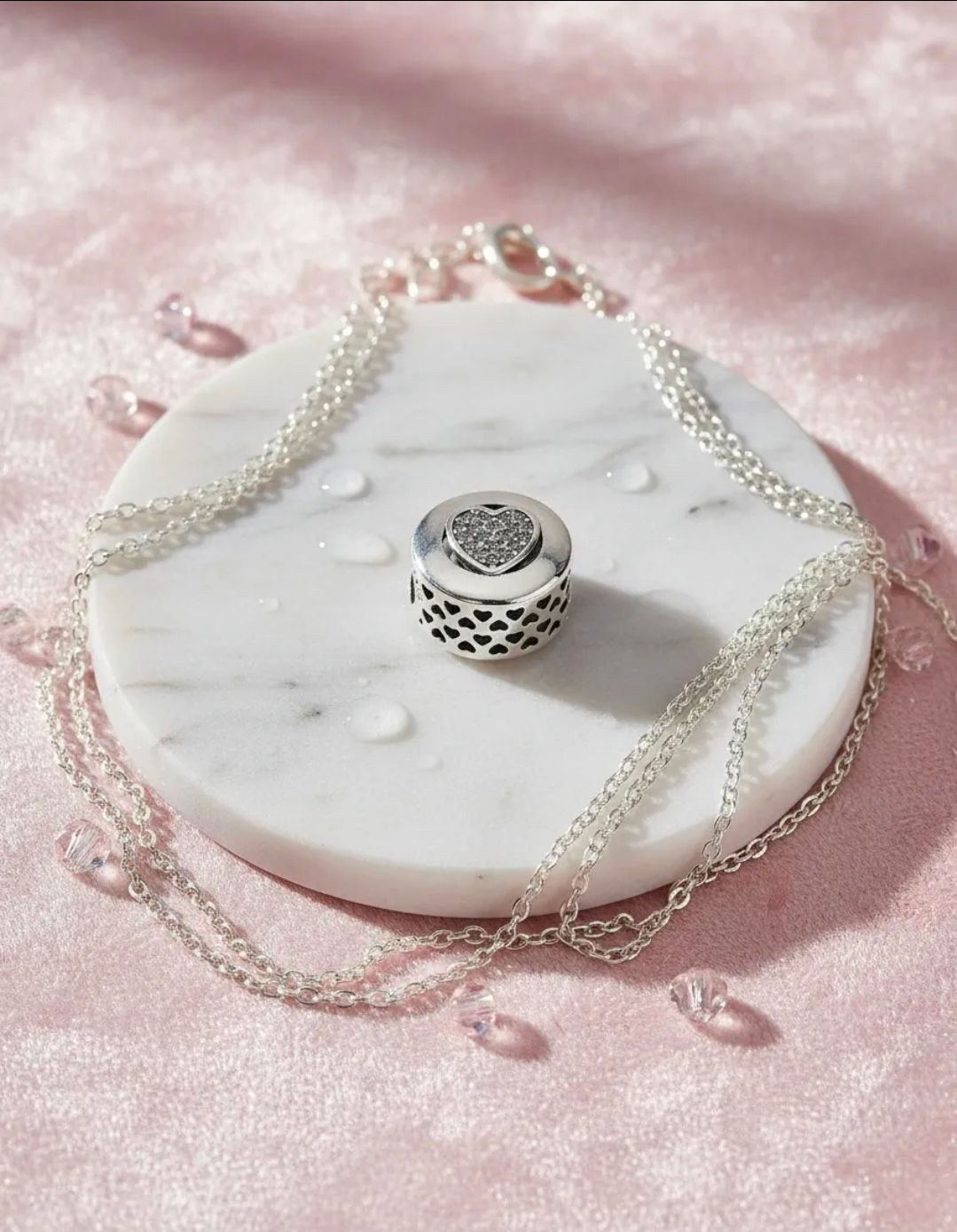 Amuleto de plata sobre un disco de mármol blanco con cadenas de perlas sobre tela de satén rosa.