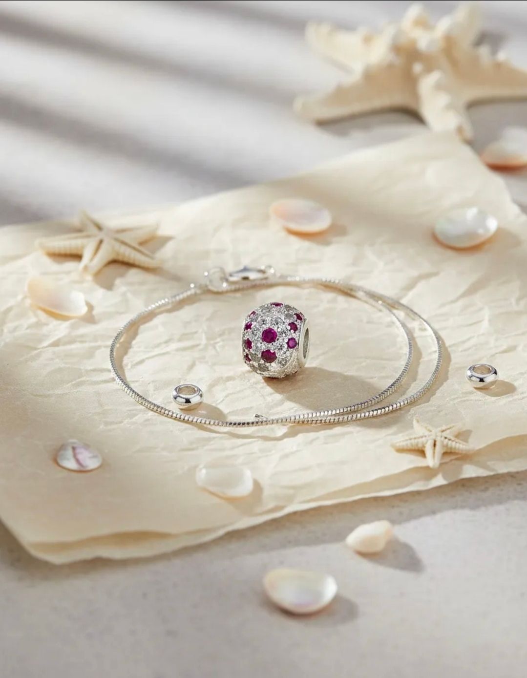 Anillo de plata con piedras preciosas rosas sobre tela beige, rodeado de conchas marinas y estrellas de mar.