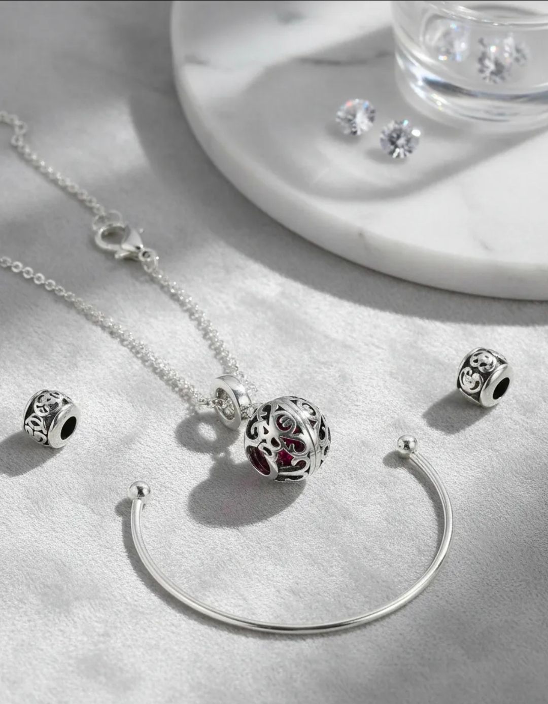 Pulsera, collar y pendientes de plata con dijes, exhibidos sobre una superficie blanca con piedras rojas y transparentes.