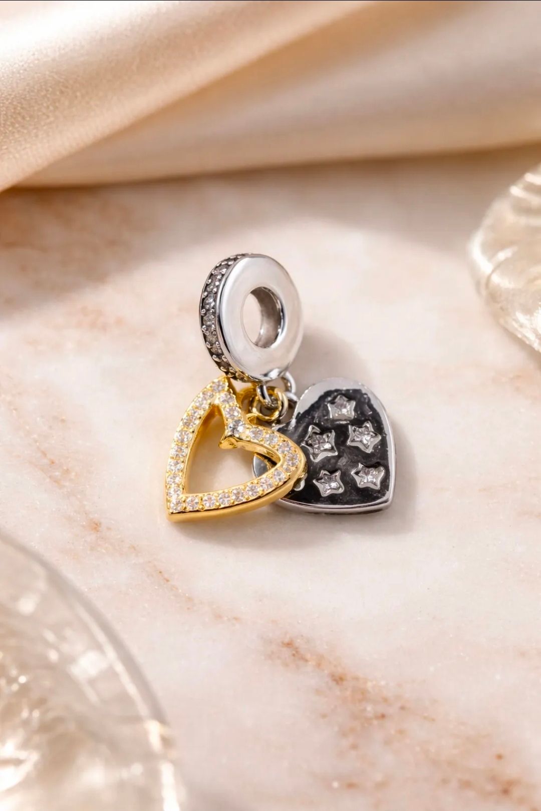 Dos colgantes en forma de corazón, uno dorado y otro negro con piedras plateadas, sobre un fondo de tela beige suave.