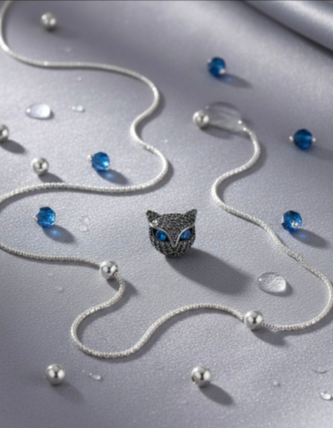 Collar de plata con detalles de cuentas azules y transparentes, con un colgante en forma de cabeza de gato sobre fondo gris.