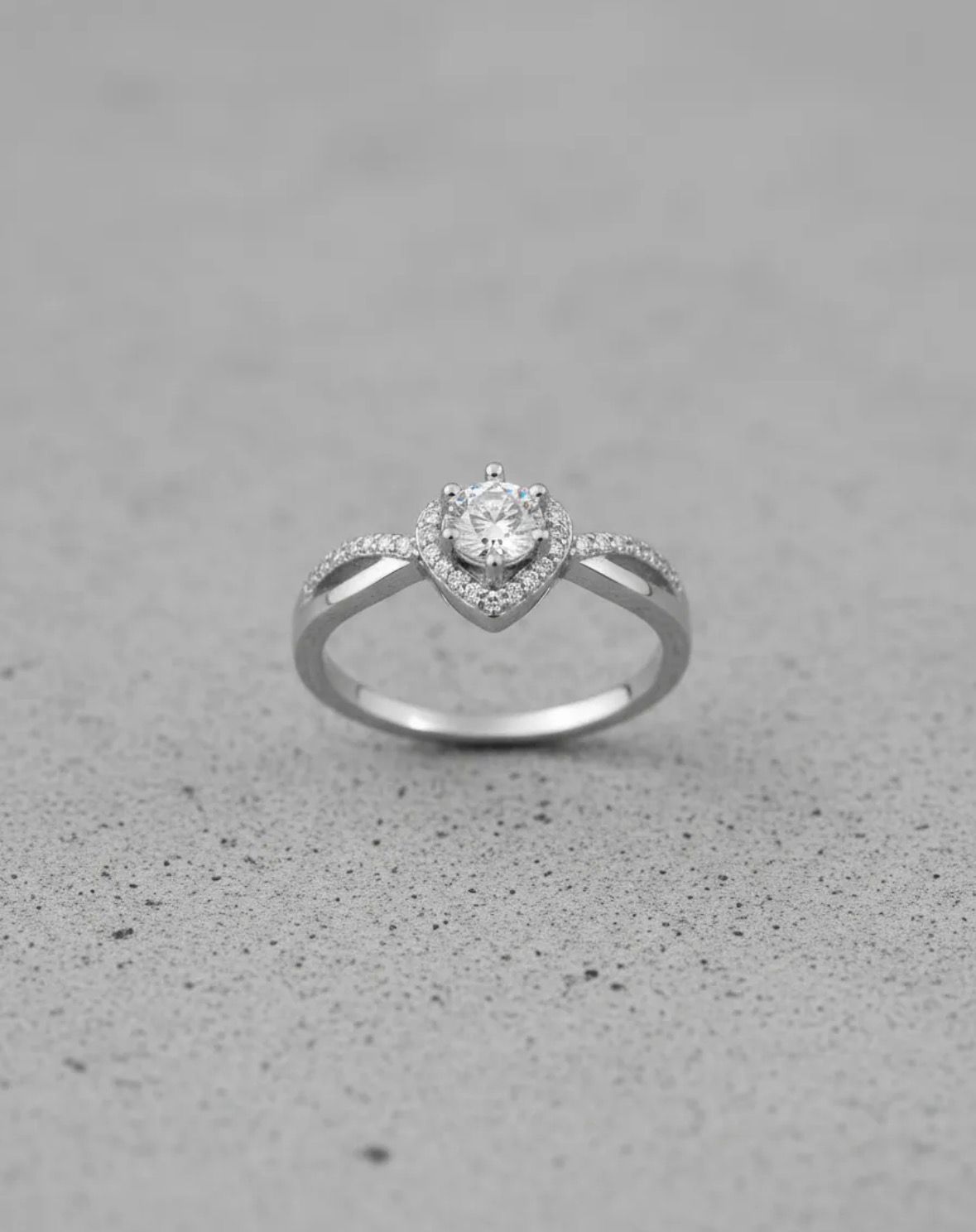 Anillo de compromiso de plata con diamante redondo sobre una superficie gris.