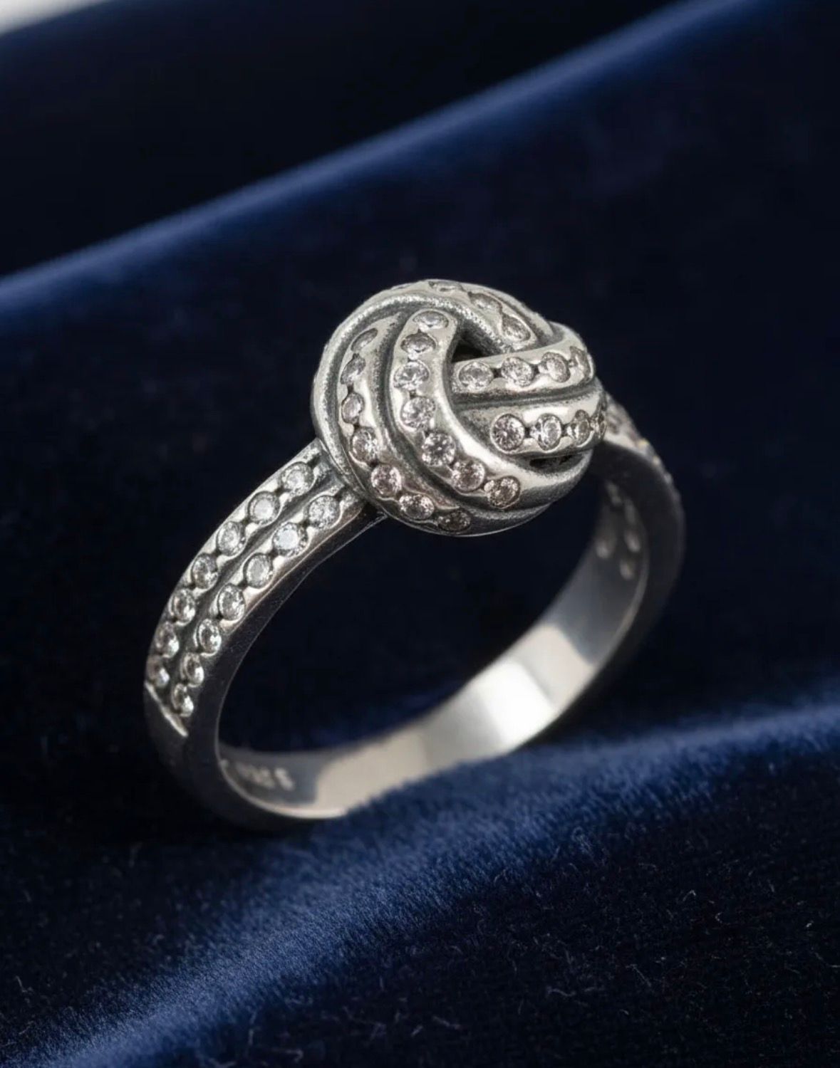 Anillo de plata con forma de nudo y pequeñas piedras transparentes sobre un fondo moteado claro.