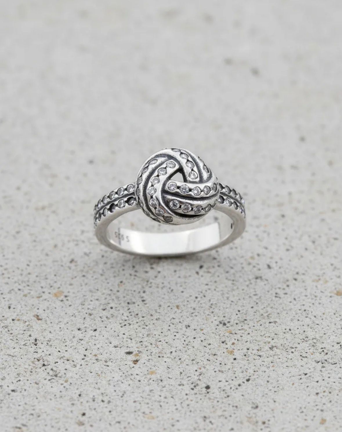 Anillo de plata con forma de nudo y pequeñas piedras transparentes sobre un fondo moteado claro.