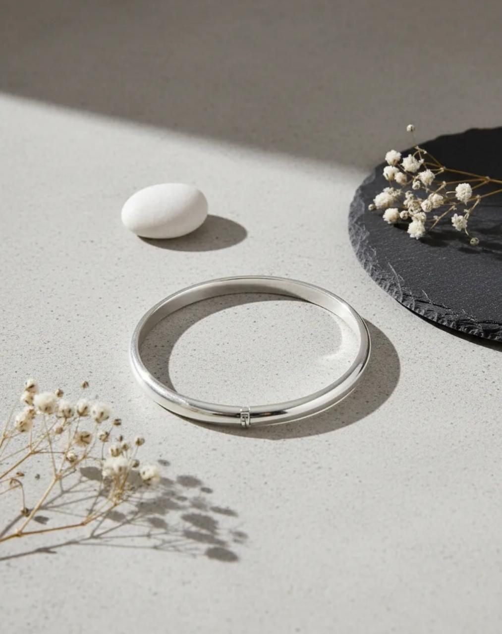 Pulsera de plata sobre una superficie clara con flores secas y una decoración de piedra blanca.
