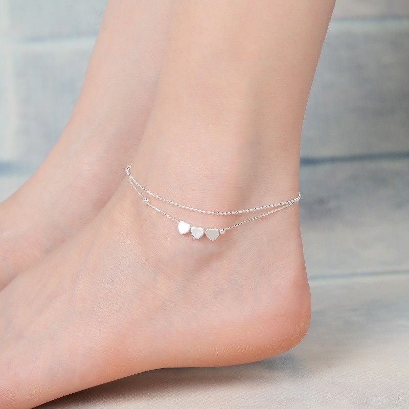 Pulsera de tobillo plateada con tres pequeños dijes de corazón en un pie descalzo