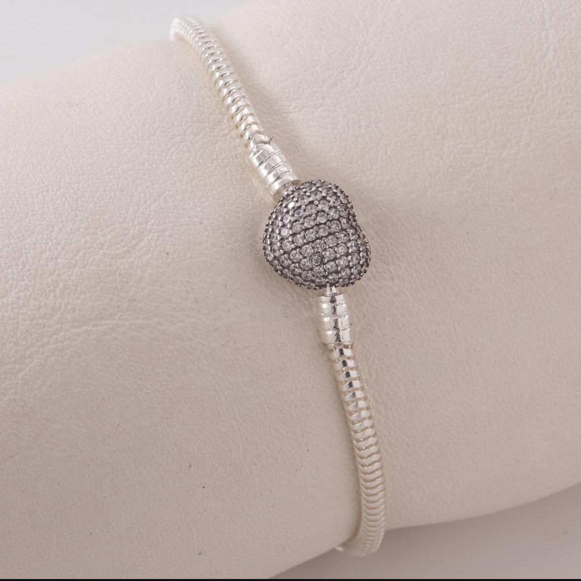 Pulsera de cuentas de plata con un dije redondo oscuro sobre una superficie acolchada de color beige.