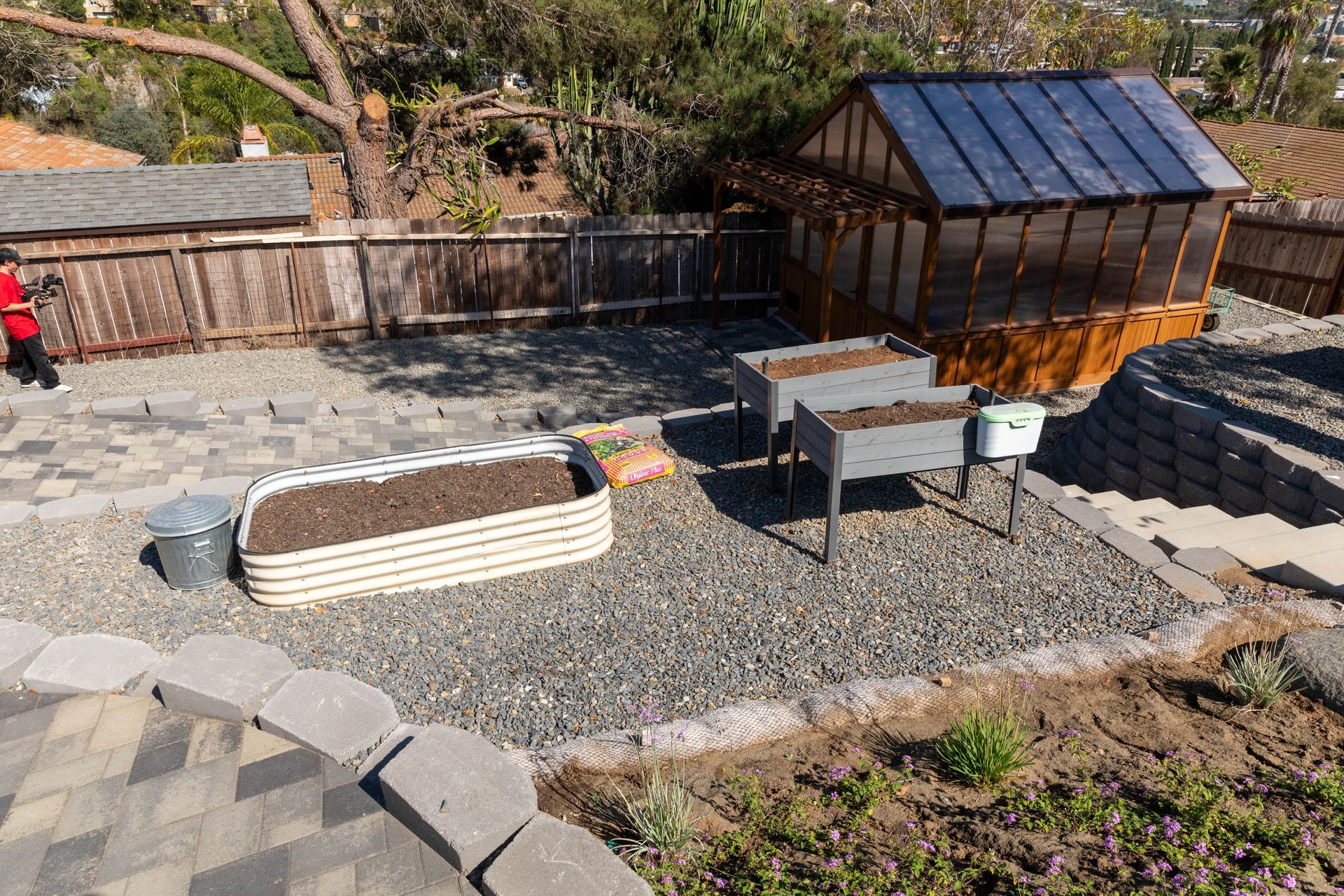 Backyard gravel patio with raised planter, small greenhouse, table, chairs, and stone steps.