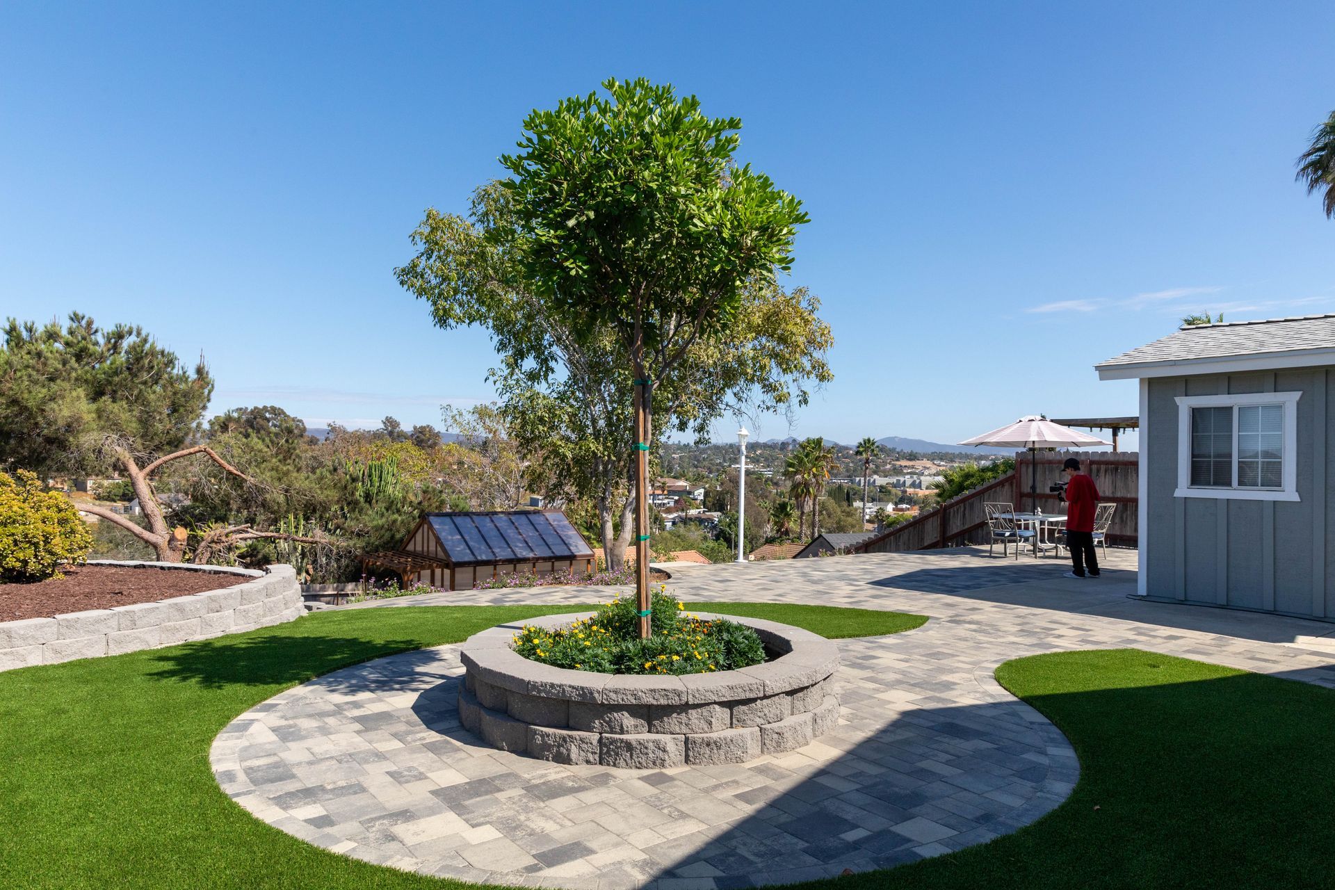 Sunny garden with circular stone planter, young tree, solar panels, and a small building beside a lawn