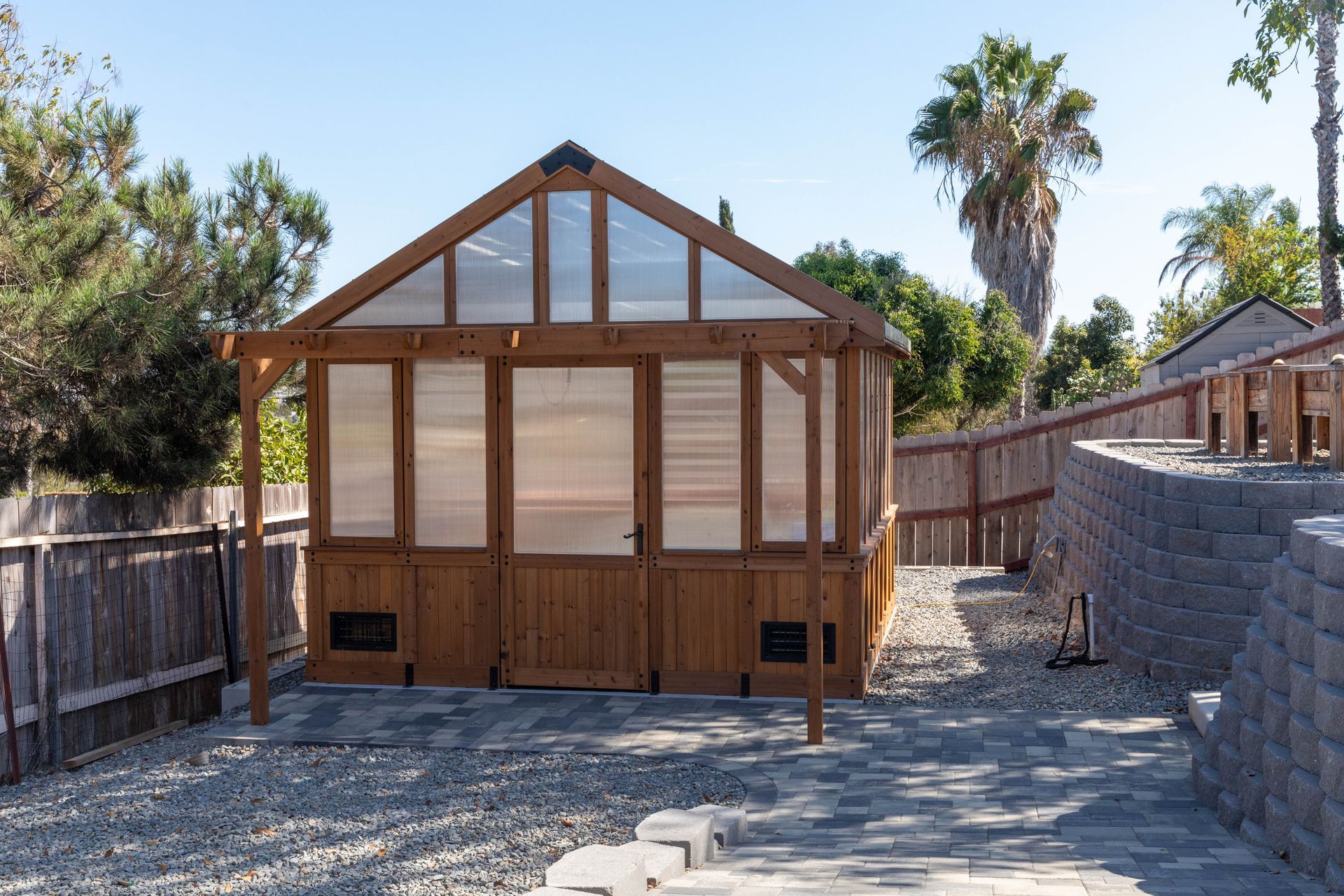 Wooden greenhouse with glass roof panels in a gravel backyard, bordered by a fence and stone retaining wall.