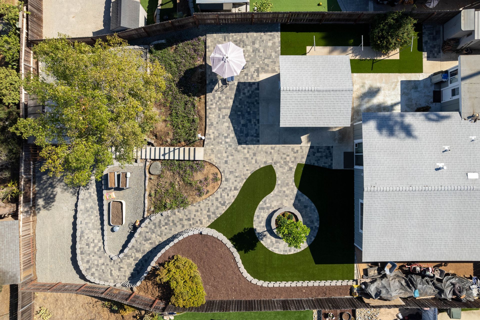 Aerial view of a landscaped backyard with curving paths, garden beds, patio seating, and a large house roof.