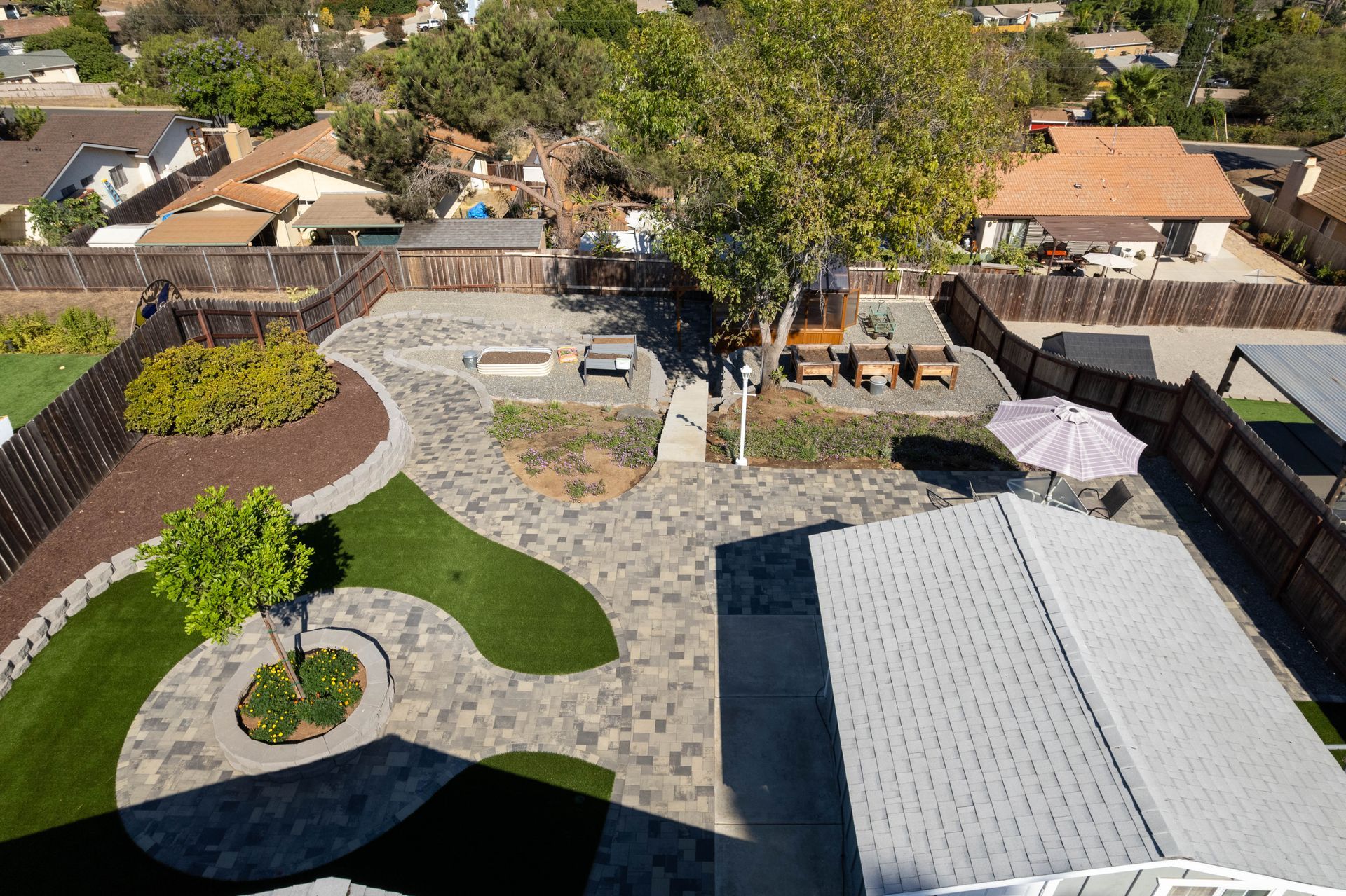Aerial view of a backyard with stone patio, green lawn, trees, and a white-roofed structure.
