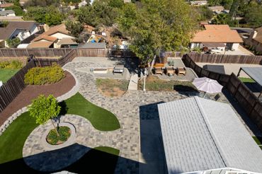 Aerial view of a backyard with stone patio, curved lawn beds, trees, and a covered patio roof.
