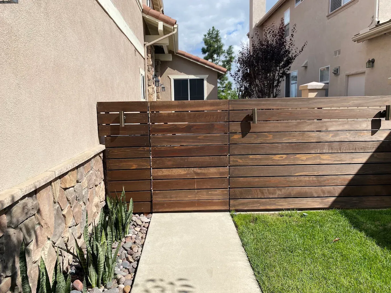 Narrow backyard walkway beside a house, with a wooden fence and green lawn under a sunny sky