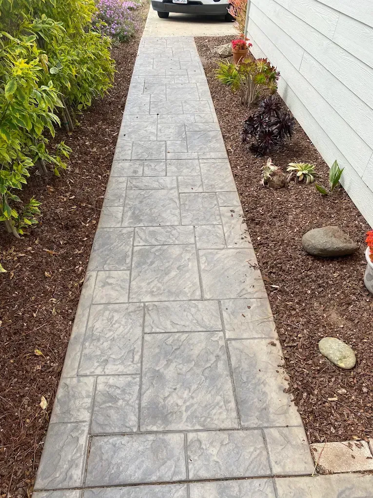 Narrow stone walkway between a hedge and house, with mulch beds and a parked car at the end.