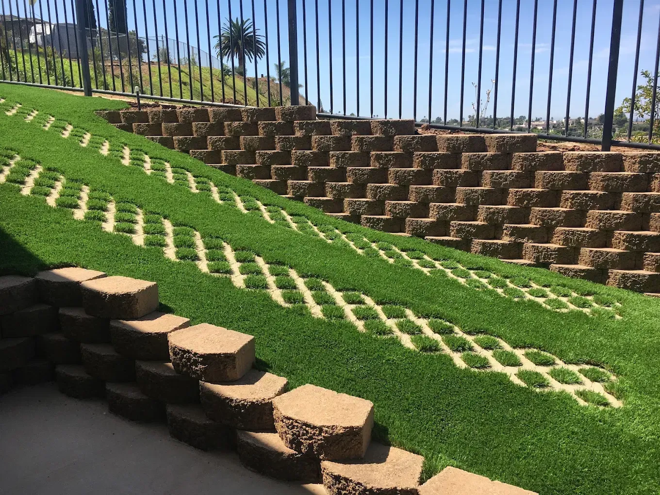 Terraced hillside with green grass, stone steps, and a black fence under a clear blue sky
