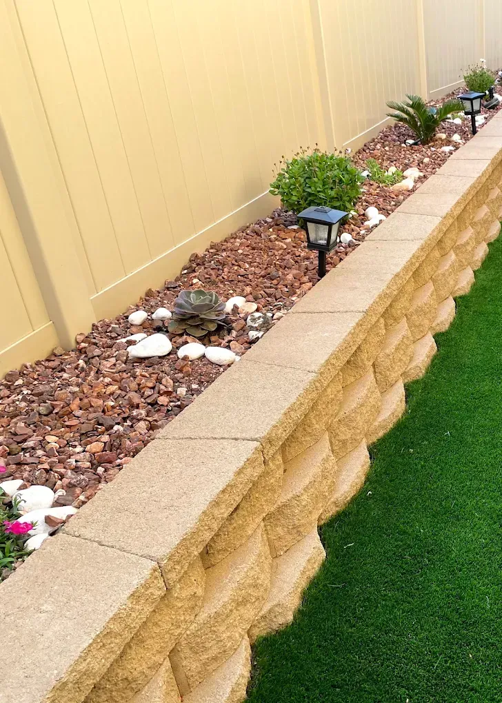 Landscaped garden bed with rocks, small plants, and a tan retaining wall beside green turf