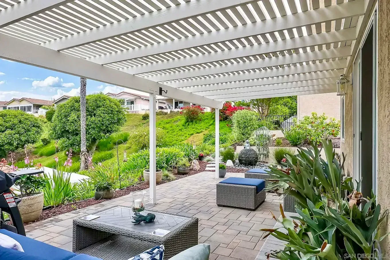 Covered patio with white pergola, wicker seating, and lush garden landscaping beyond a brick walkway