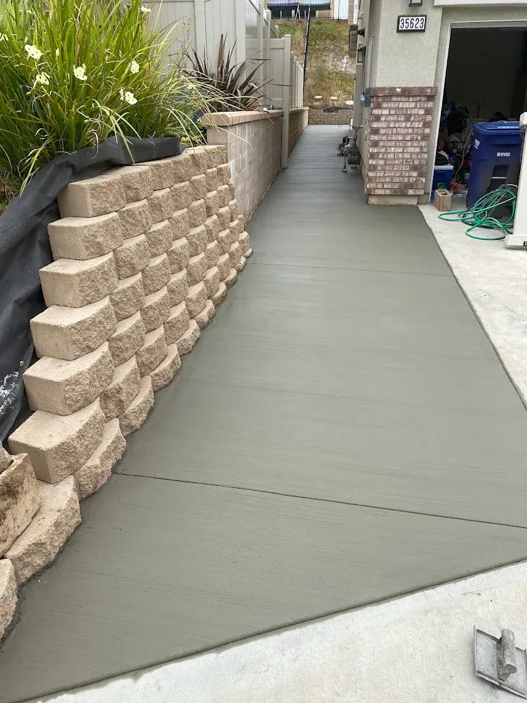 Concrete driveway beside a brick house, with a retaining wall and landscaped plants on the left.