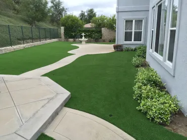 Backyard patio with artificial turf, curved walkway, and a house wall with windows and shrubs