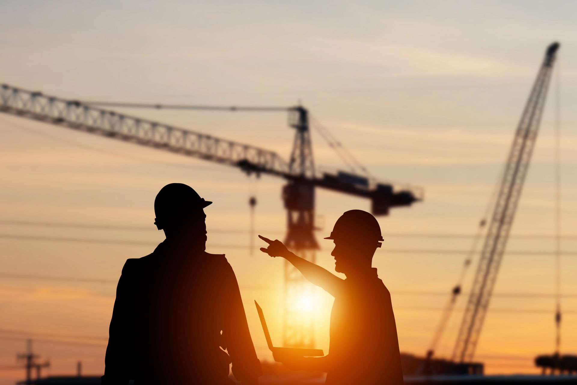 Operai edili in silhouette con caschi protettivi, gru e computer portatile al tramonto. Uno indica il cantiere.