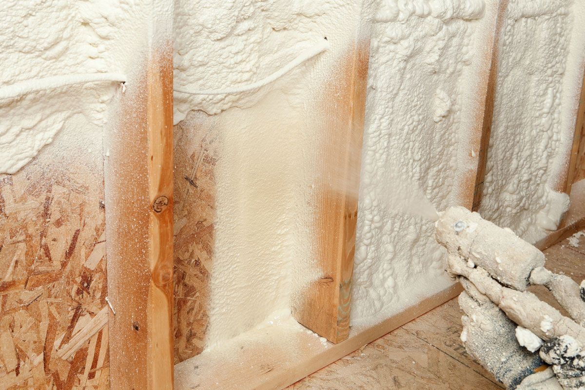 A spray gun applies white foam insulation into the wooden stud wall of a building under construction.