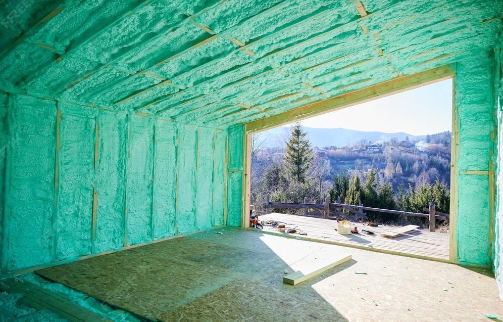 A room under construction with turquoise spray foam insulation on the walls and ceiling, looking out to a mountain view.