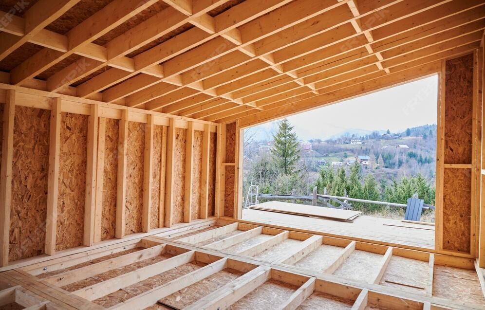The interior of an unfinished wooden building structure with exposed wall studs, floor joists, and a view of mountains.