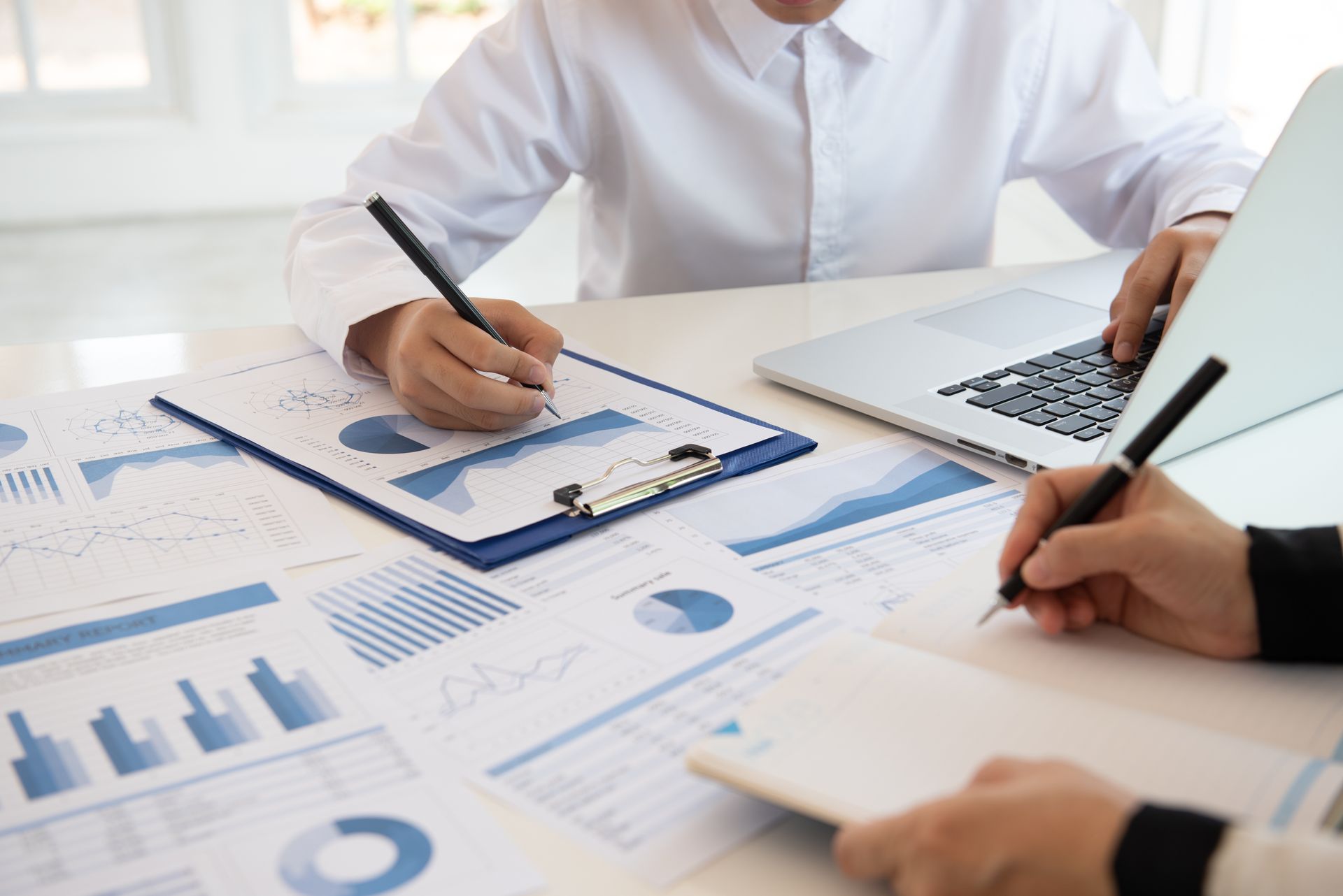 Two people sitting at a desk reviewing financial documents and charts, one using a laptop and both taking notes.