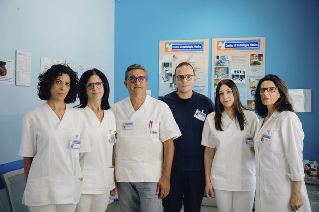Cinque membri del personale medico in camice bianco sono in piedi in una clinica con dei poster appesi al muro.