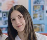 Persona con lunghi capelli castani sorridente in una stanza luminosa con una bacheca sfocata sullo sfondo.