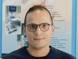 Ritratto di una persona con occhiali rotondi e camicia scura, in piedi davanti a uno sfondo blu da poster.
