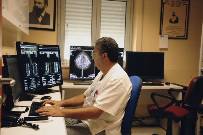 Un medico esamina le scansioni cerebrali su due monitor in un ufficio con poster medici e una scrivania.