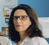Donna con capelli scuri e occhiali, in camice bianco, in piedi in un laboratorio o in un ufficio.