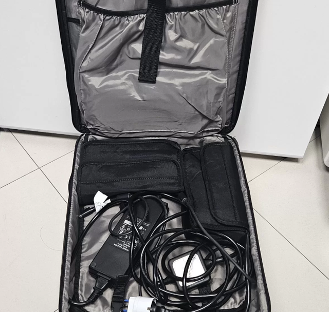 Borsa nera aperta con cavi arrotolati e caricabatterie su un pavimento piastrellato.
