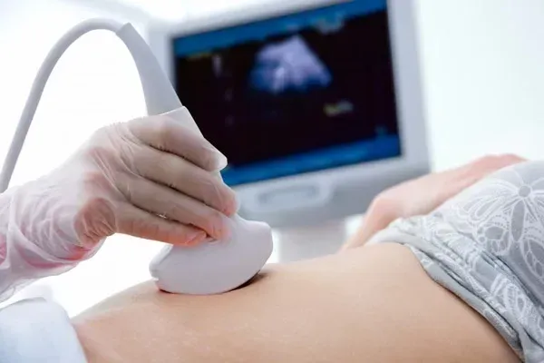 Ecografia eseguita con una mano guantata che regge una sonda sull'addome di una donna incinta, con monitor sullo sfondo.